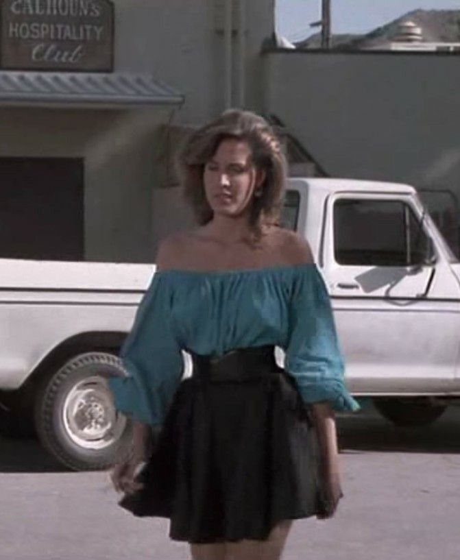 Woman wearing a teal off-the-shoulder blouse and a black skirt standing by a white truck.