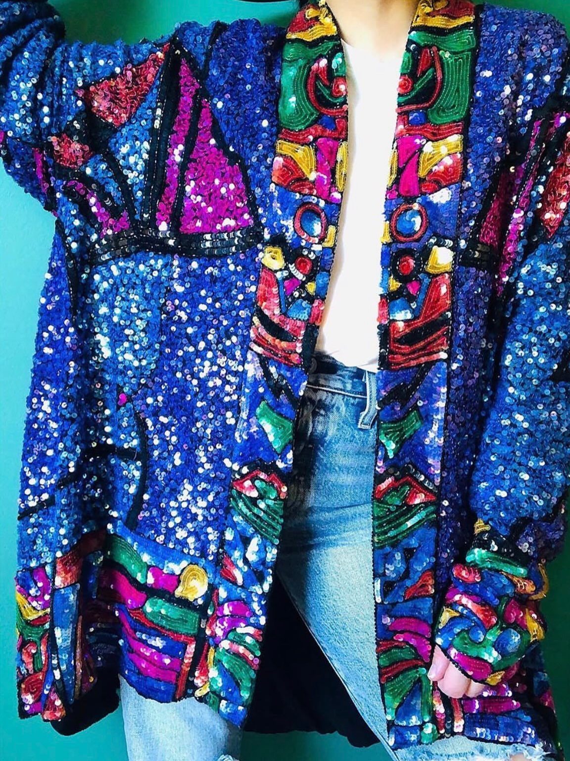 A person wearing a bright blue sequined jacket with colorful geometric patterns and denim jeans.