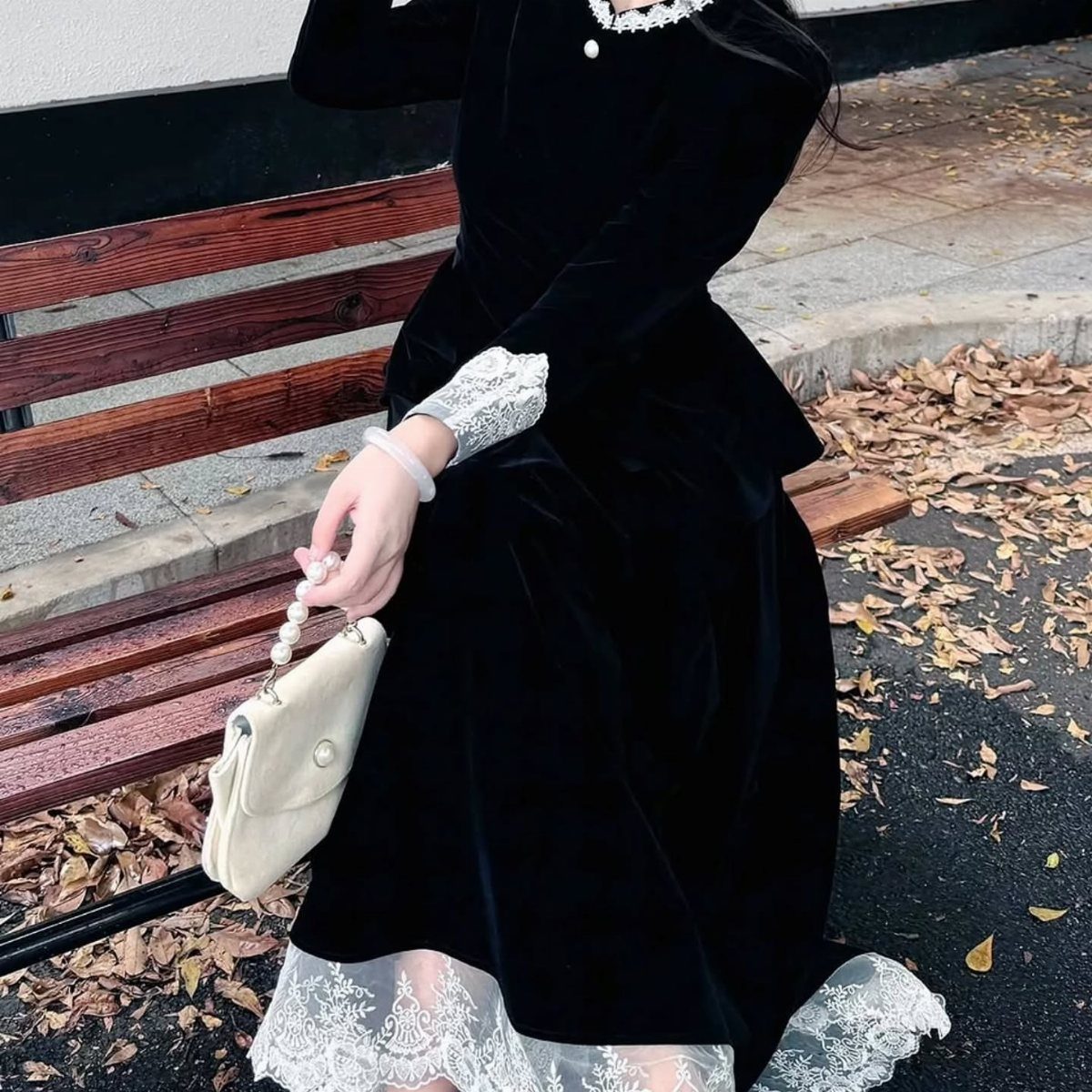 A person wearing a long-sleeved black velvet dress with white lace detailing sitting on a bench.