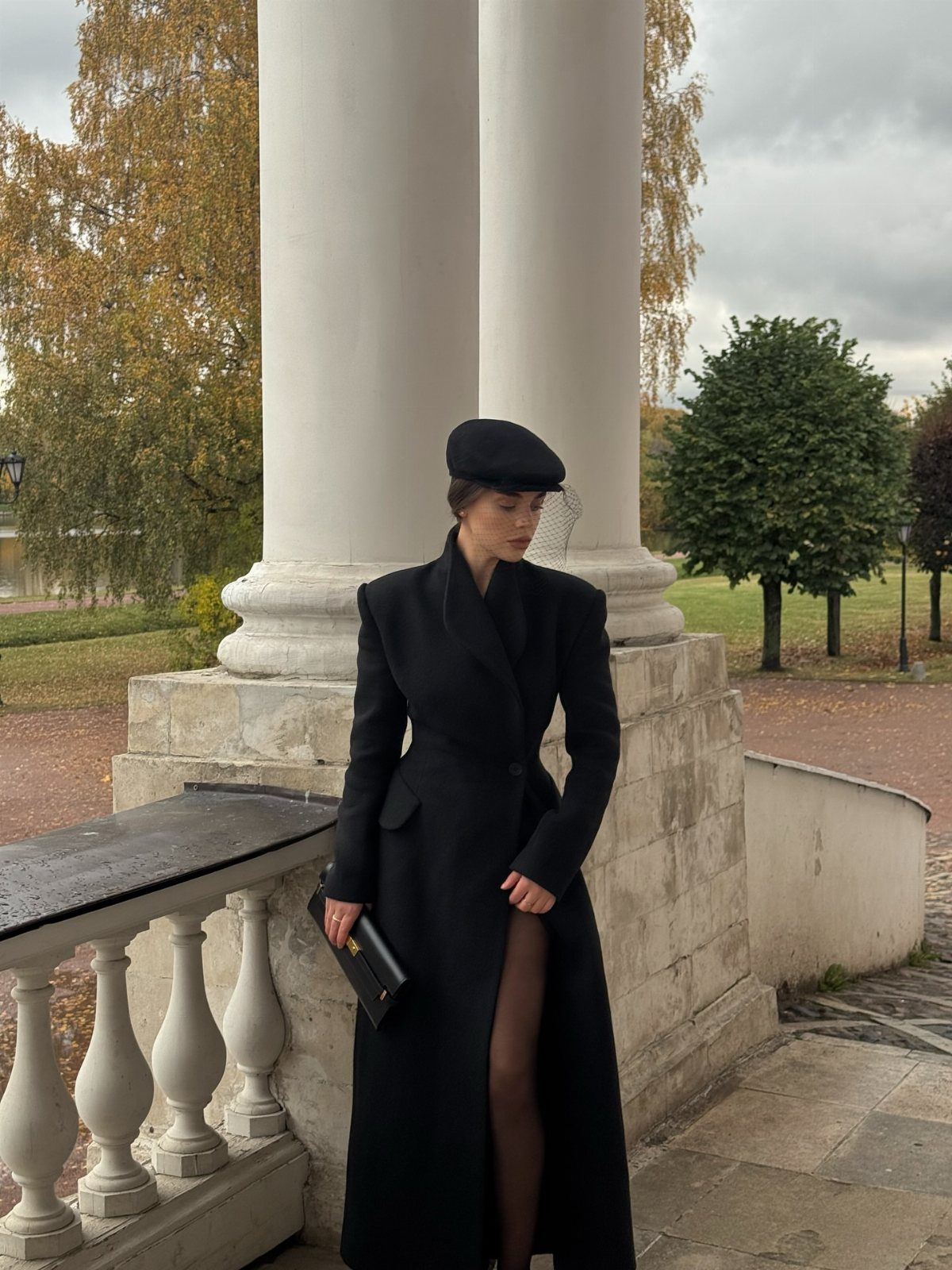 A woman wearing a long black coat and beret standing by white stone columns.