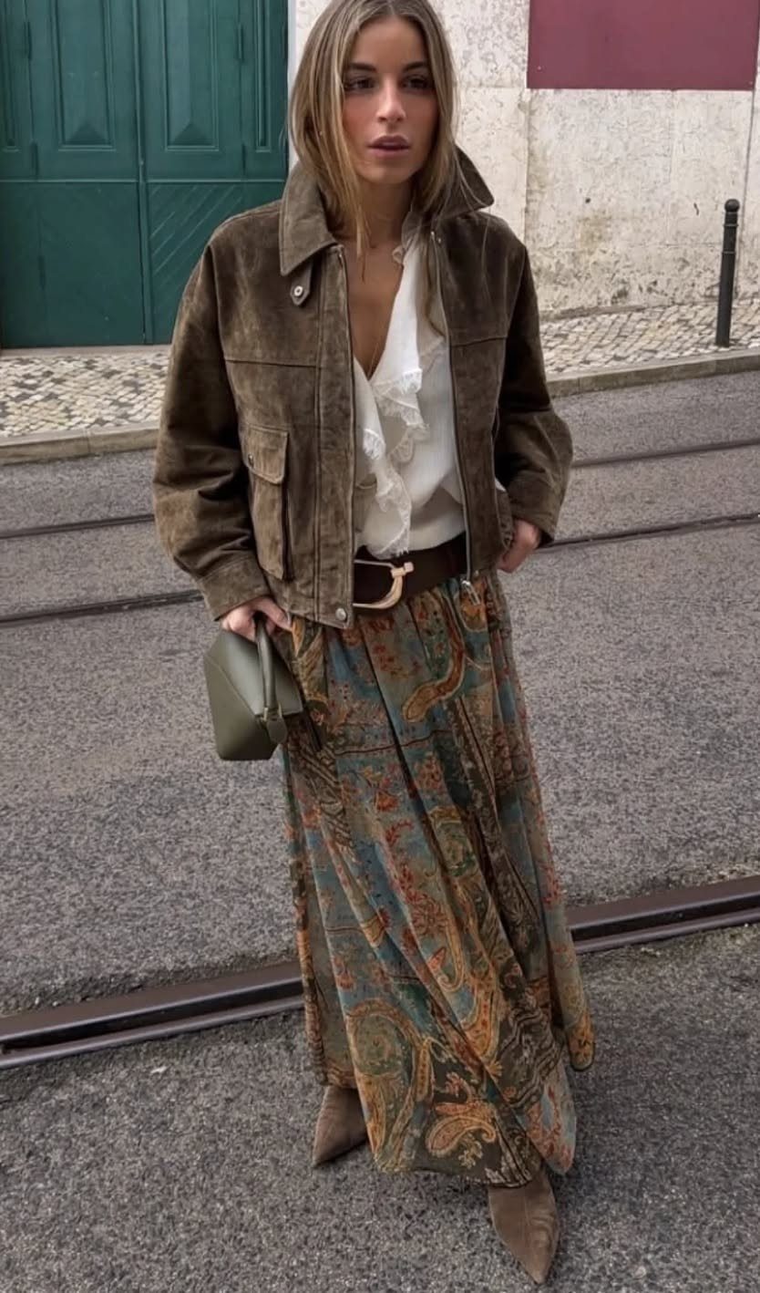 Woman wearing a brown suede jacket, white ruffled blouse, and a long patterned bohemian maxi skirt.