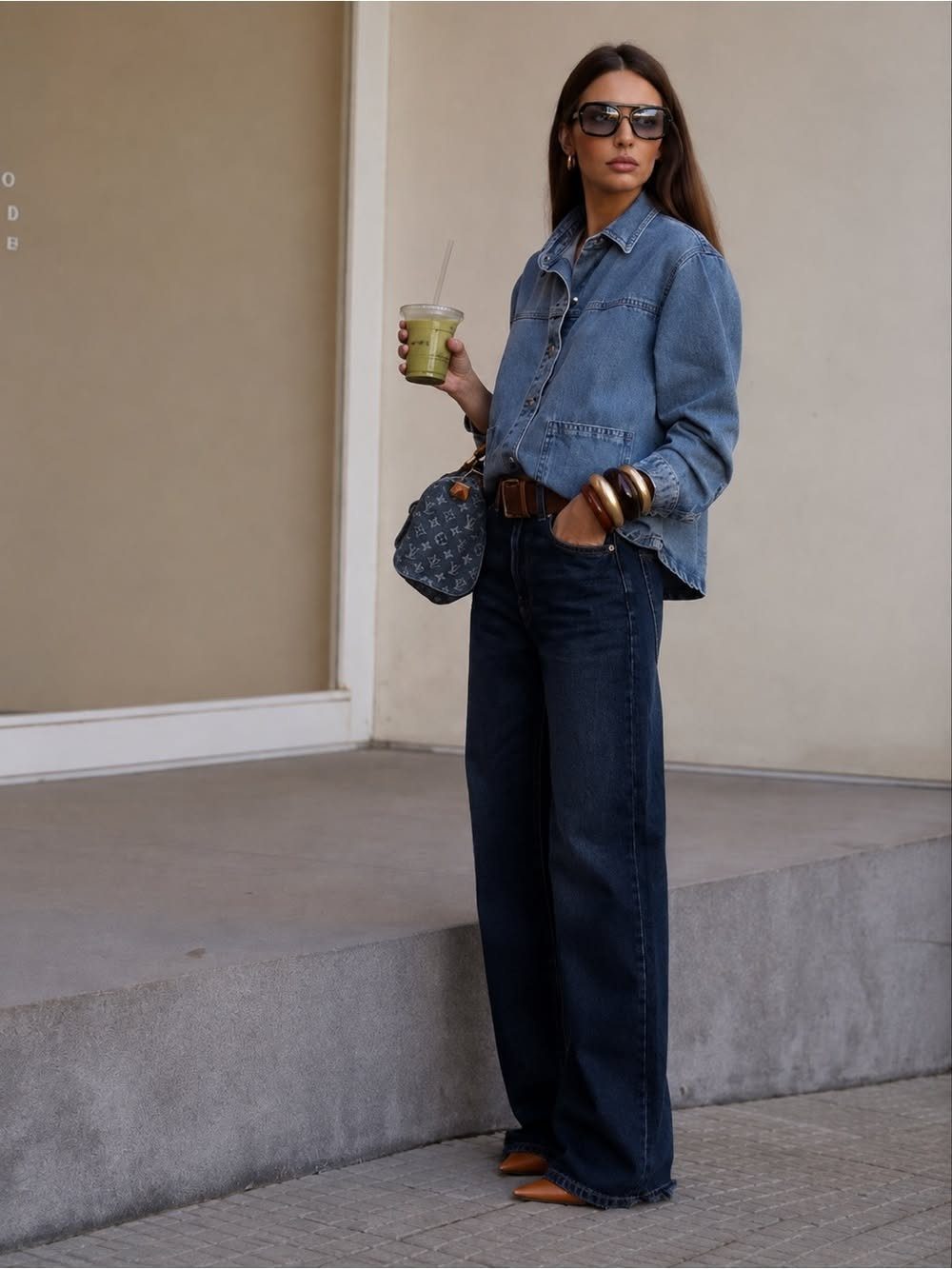 Woman wearing a denim shirt and dark wide-leg jeans standing outdoors with a drink.