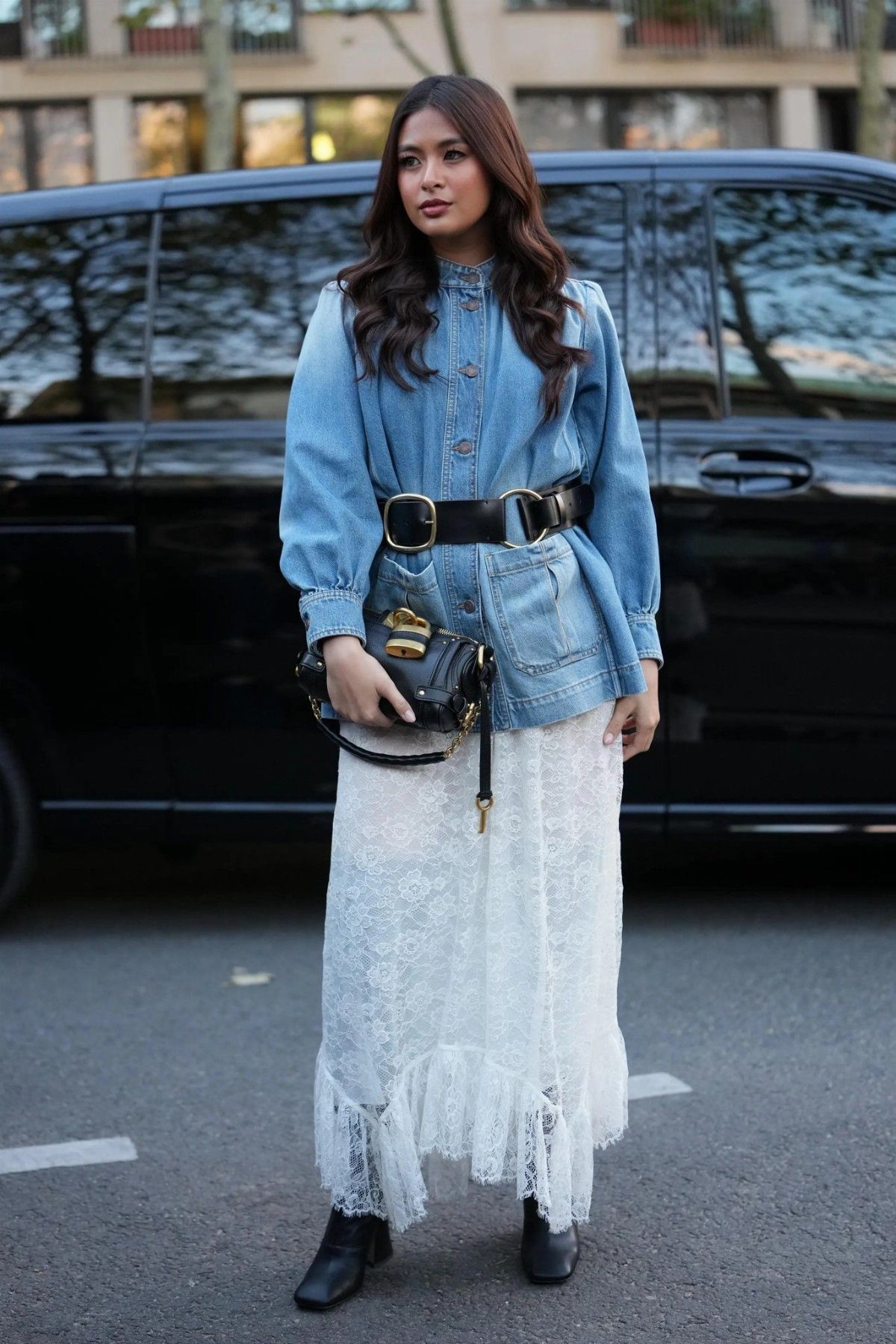A woman wearing a blue denim jacket and a long white lace skirt standing on a street.