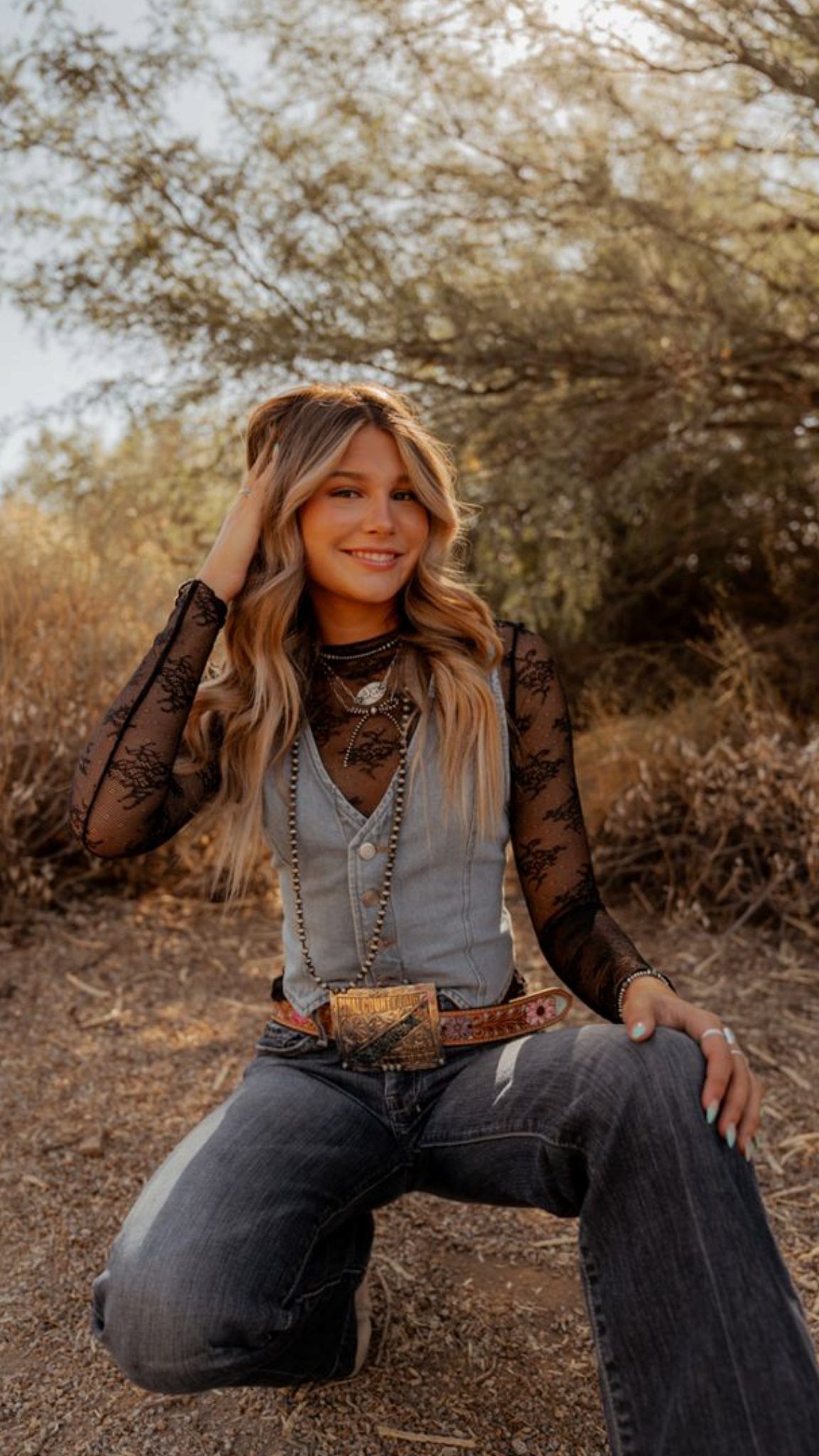 A smiling woman wearing a lace top, denim vest, and dark jeans posing in a field.