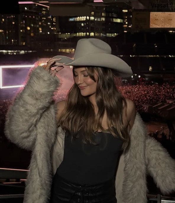 Woman in a white cowboy hat, dark corset, and fluffy white coat smiling at night.
