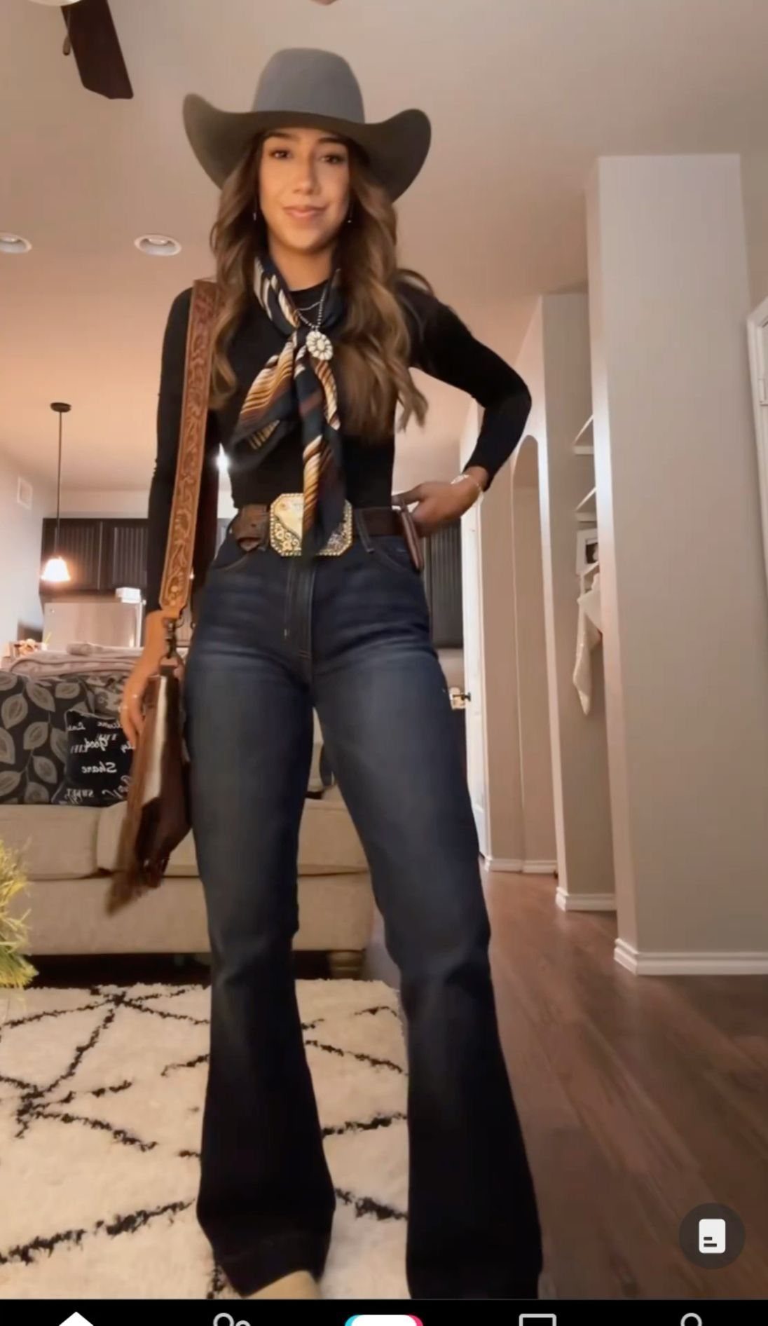 Woman wearing flared denim jeans, a black top, large western belt, and a grey cowboy hat.