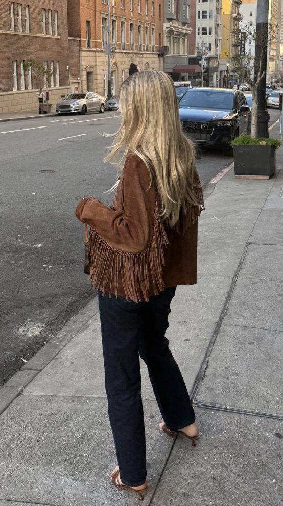 Blonde woman walking on a city sidewalk wearing a brown suede fringe jacket and dark denim jeans.