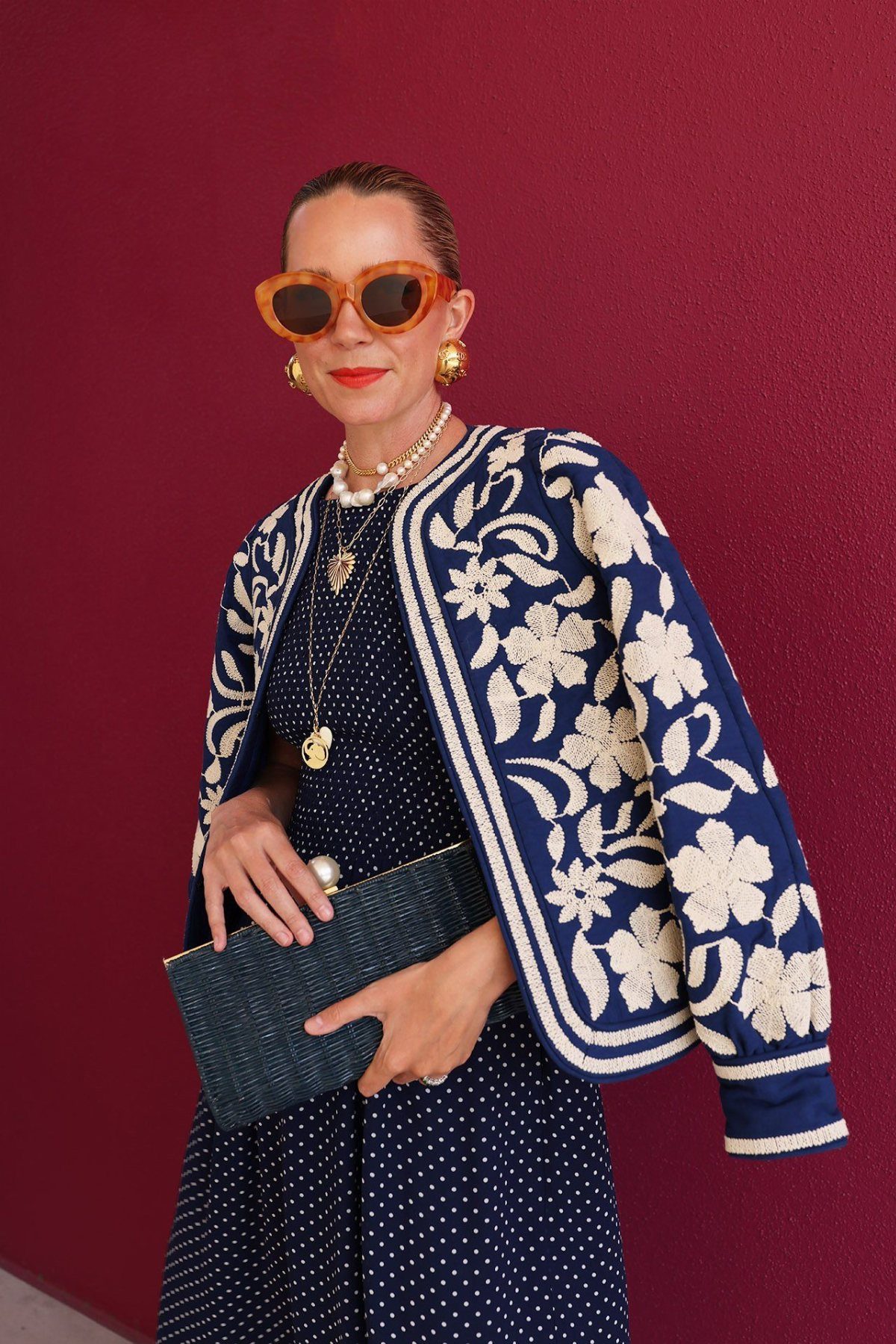 Woman wearing a navy polka dot dress and a cream floral embroidered jacket against a burgundy wall.