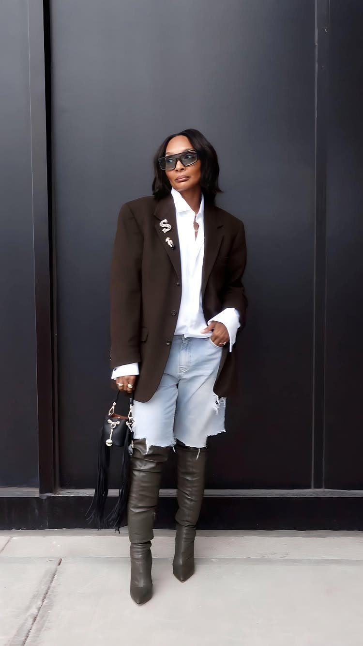 A woman in a brown blazer, denim shorts, and tall green boots posing against a dark wall.
