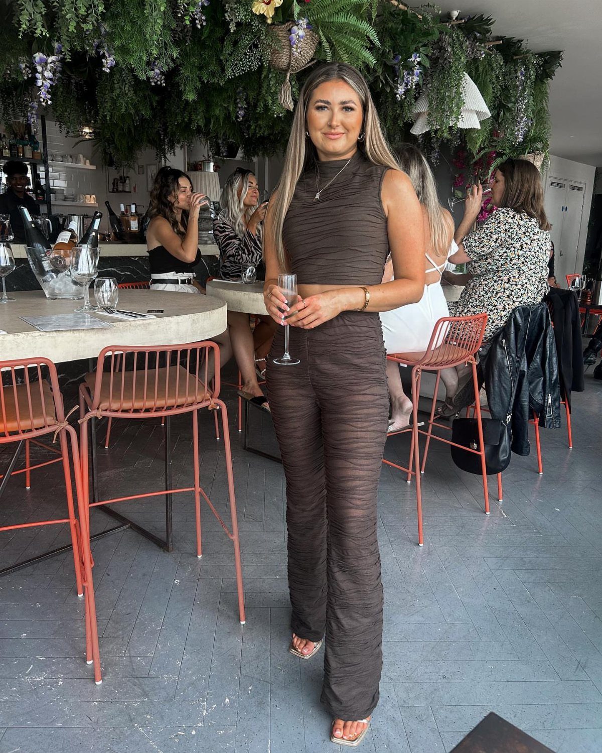 A woman in a brown textured two-piece outfit holding a glass in a restaurant.