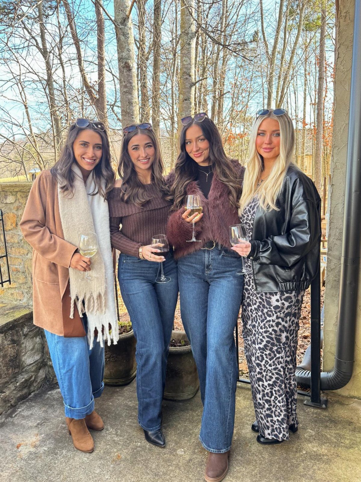 Four women smiling and holding wine glasses while wearing stylish fall layered outfits outdoors.