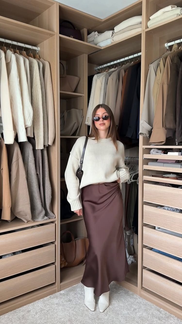 Woman in a cream knit sweater and brown satin skirt standing in a large walk-in closet.