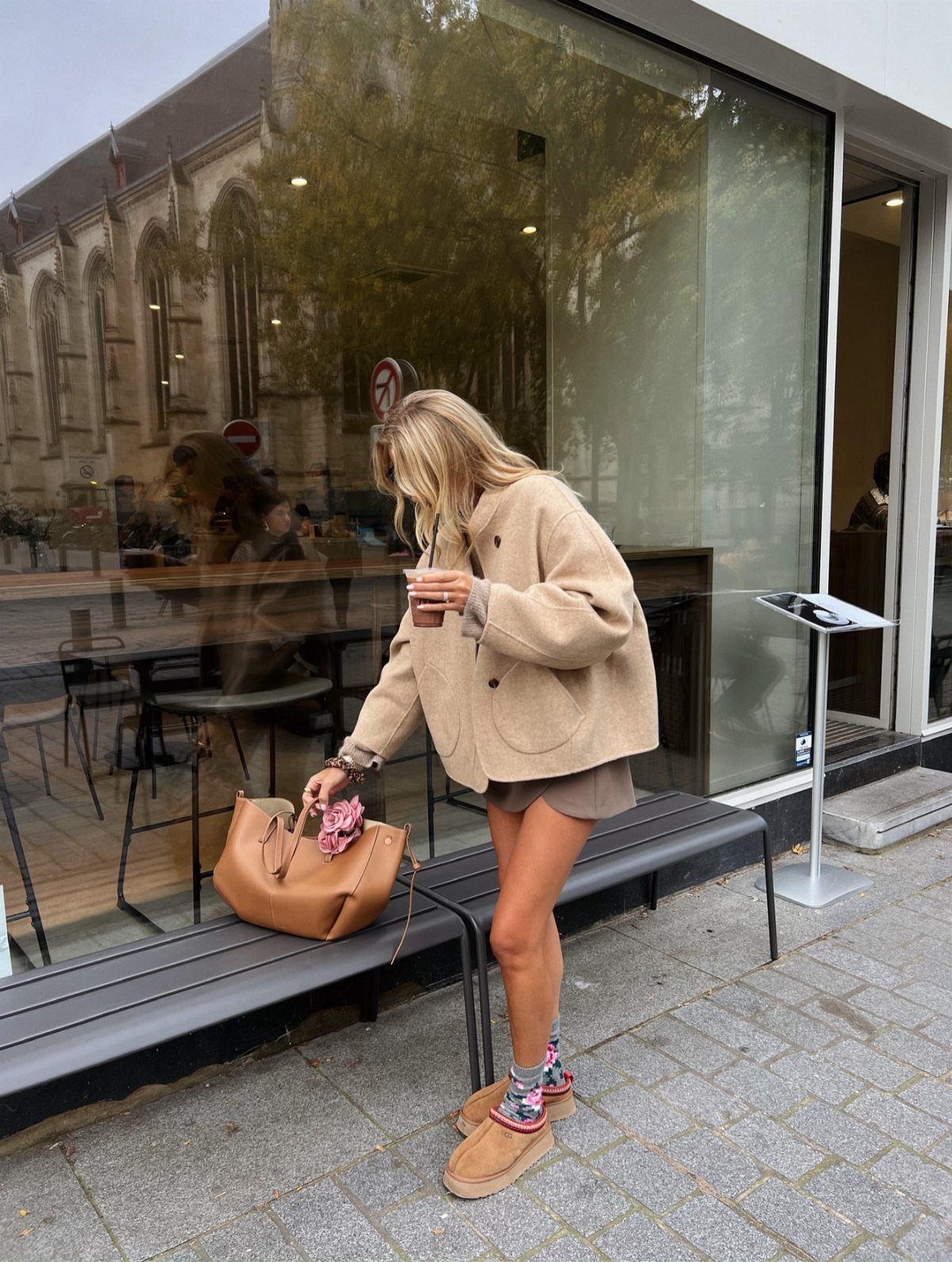 Blonde woman in a tan cardigan, tan skirt, patterned socks, and platform clogs standing by a glass window.