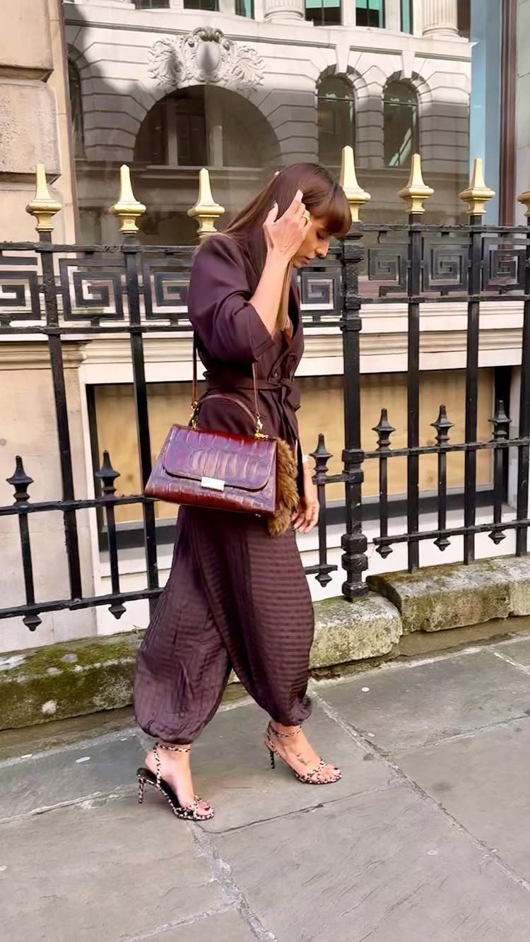 Woman in a brown jumpsuit walking with a burgundy handbag and high heels.