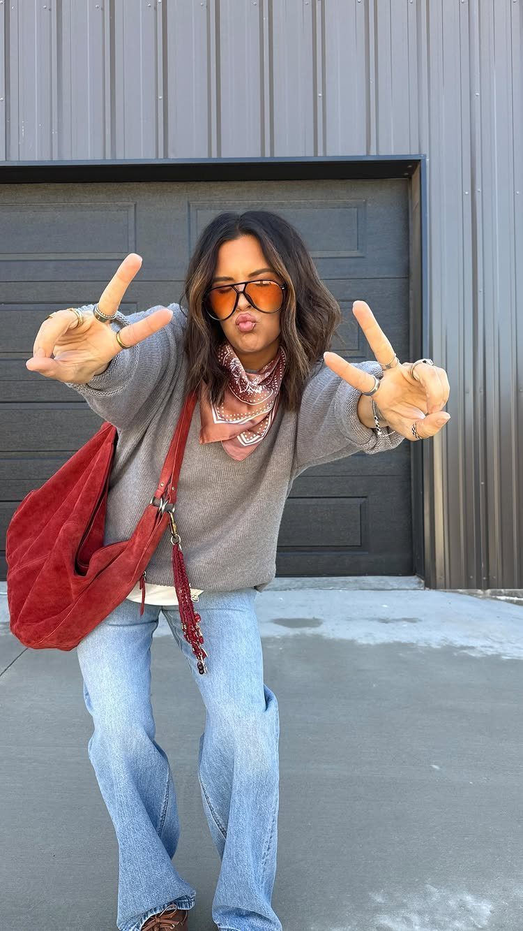 Woman in a grey sweater, blue jeans, and a pink patterned scarf posing outdoors.