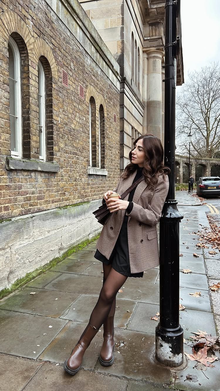 Woman in a plaid blazer, black dress, and brown ankle boots standing by a lamppost.