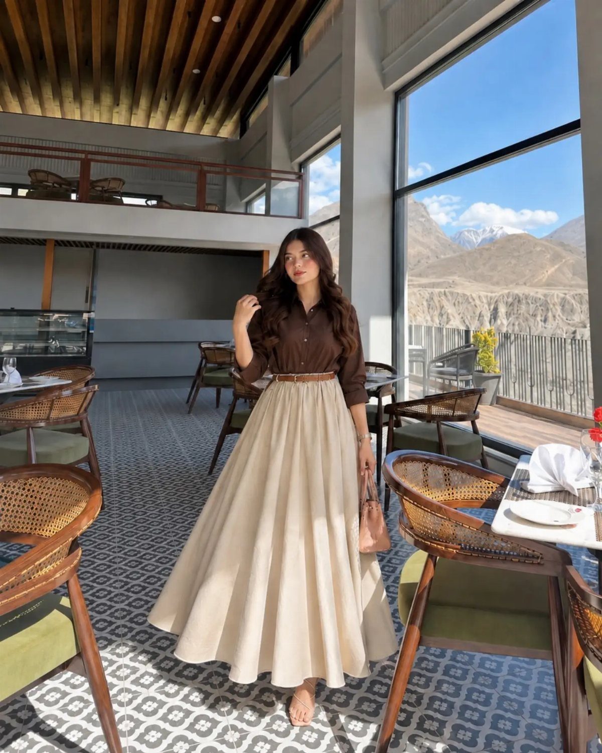 Woman wearing a dark brown button-down shirt and a voluminous cream maxi skirt standing in a restaurant.