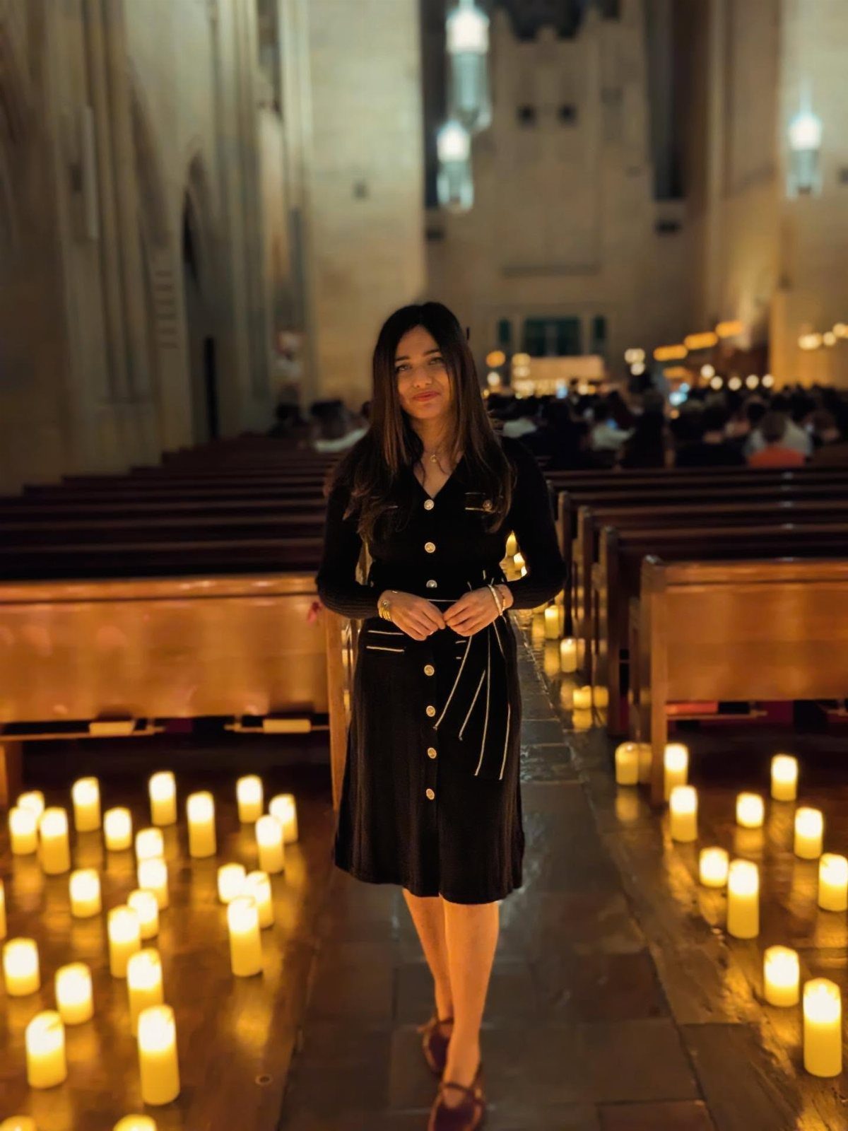 Woman in a long-sleeved black dress standing in a candlelit church aisle.