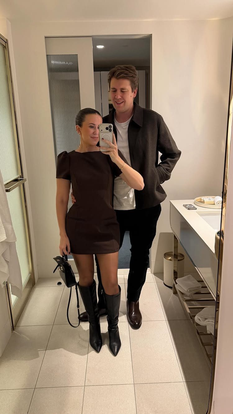 Woman in a brown mini dress and black boots posing for a mirror selfie with a man.