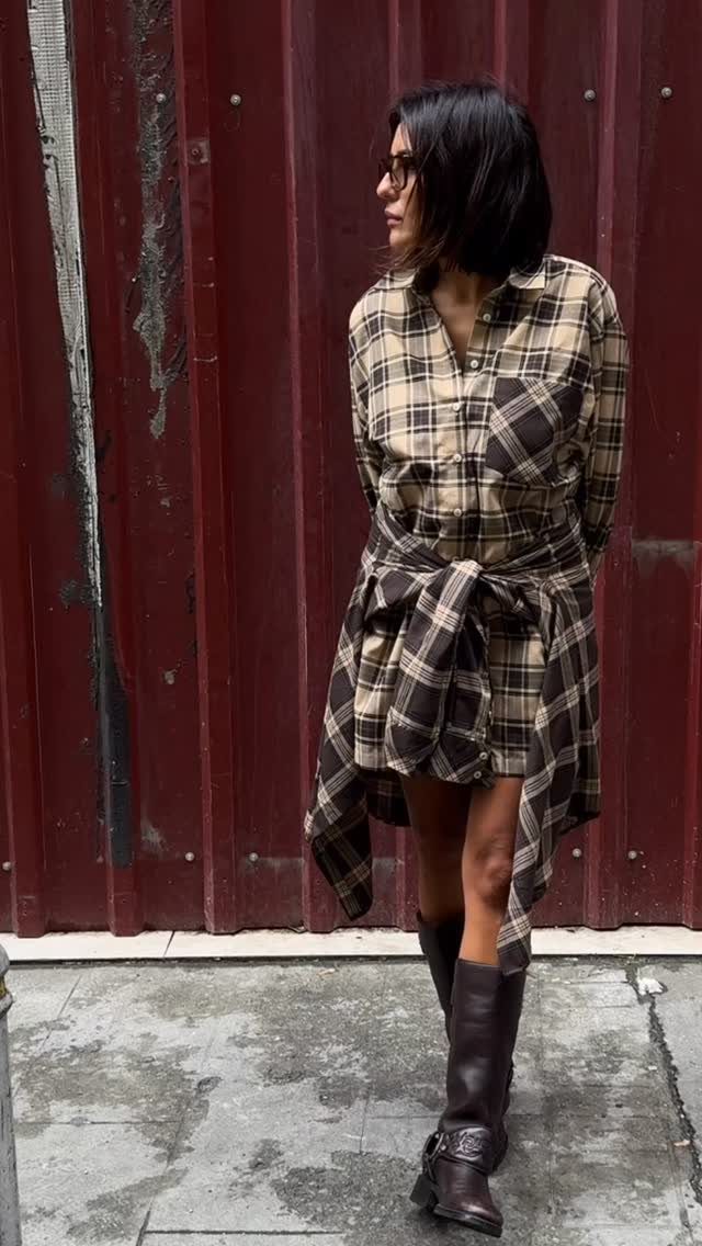 Woman wearing a brown plaid shirt dress and dark brown knee-high boots against a red wall.