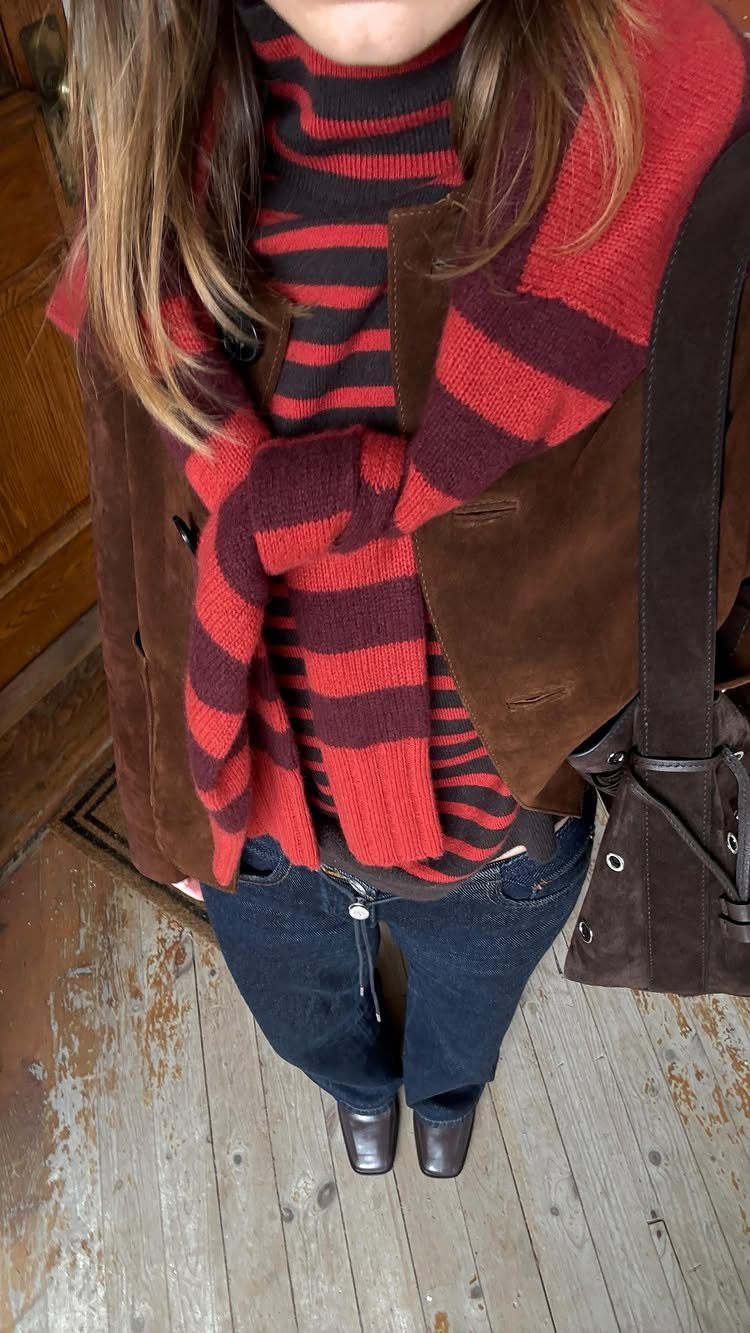 A person wearing a striped turtleneck, brown suede jacket, and dark jeans standing on a wooden floor.