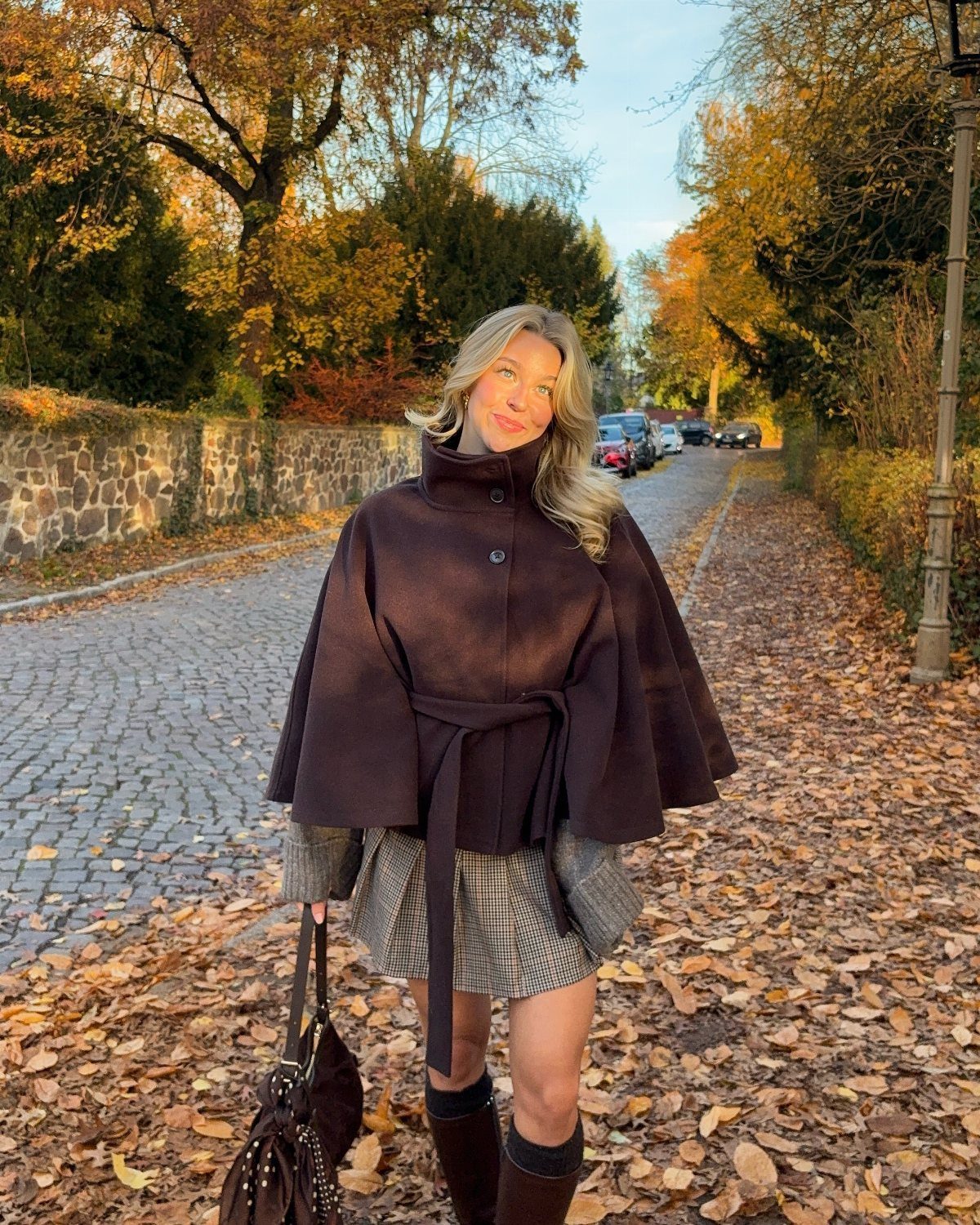 A woman wearing a dark brown belted cape jacket and a patterned skirt standing on a leaf-covered path.