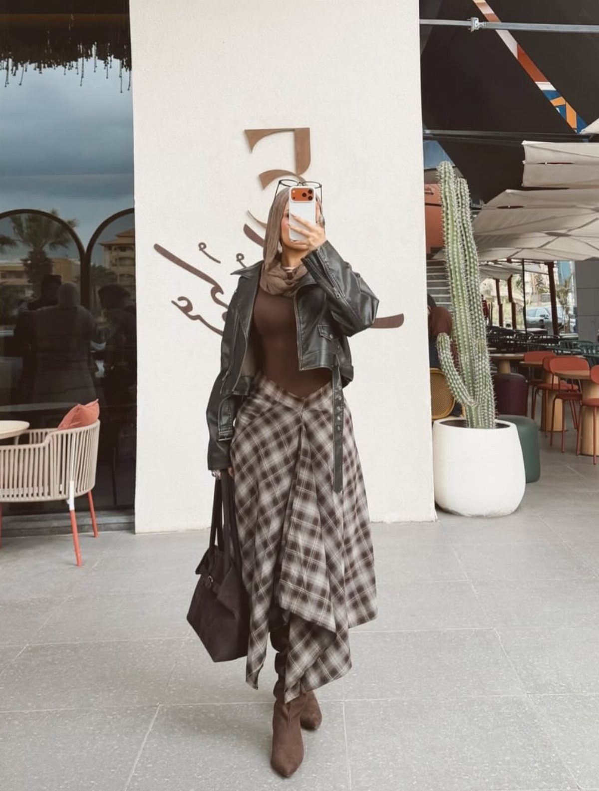 Person in a leather jacket and plaid skirt taking a mirror selfie outdoors.