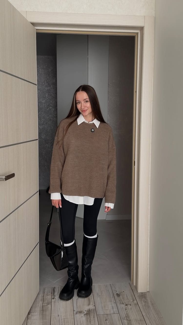 Woman wearing a brown sweater, white shirt, black leggings, and tall black boots standing in a doorway.
