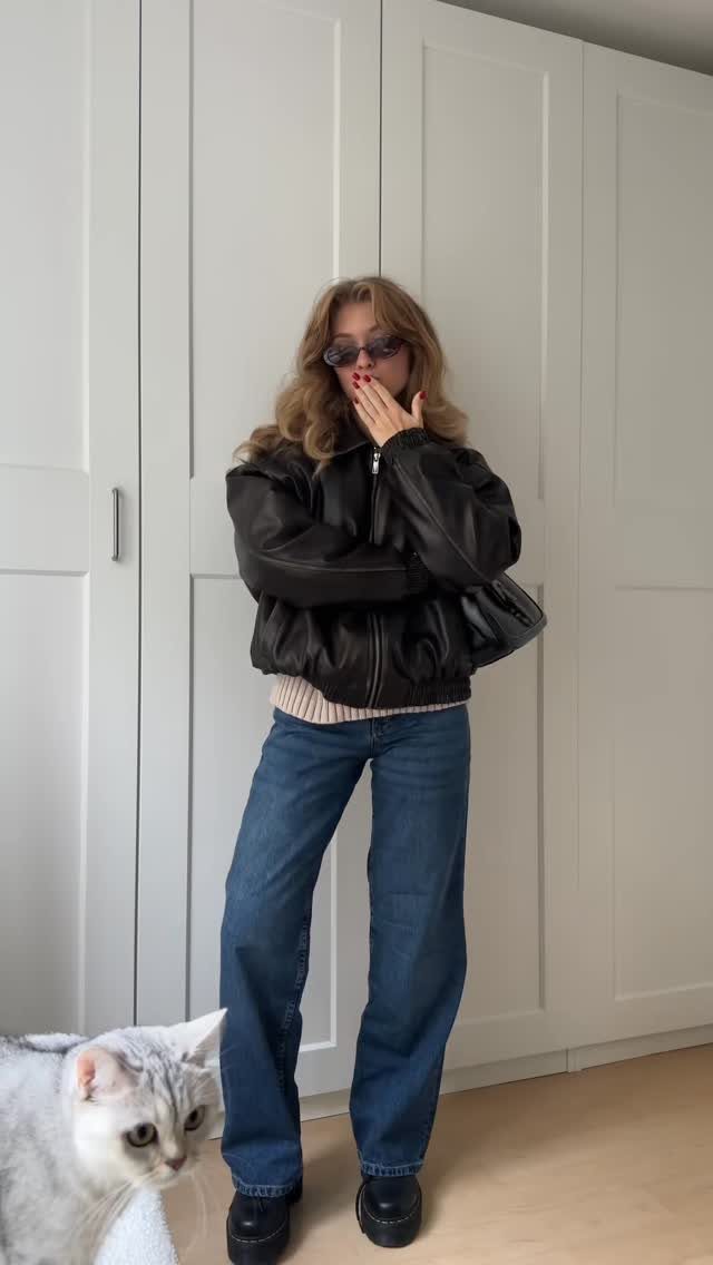 A woman wearing a black leather puffer jacket and blue denim jeans standing near a white cat.