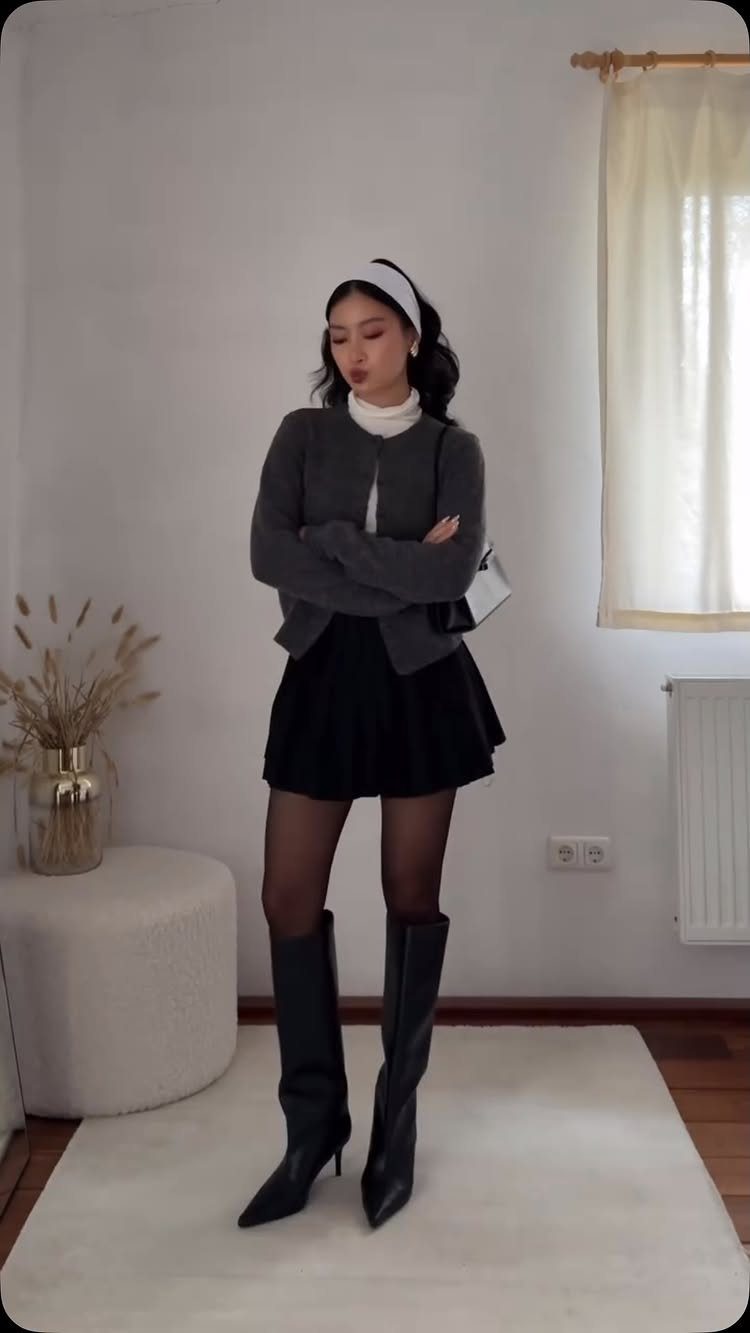 Woman wearing a grey cardigan over a white turtleneck, black skirt, and tall black boots.