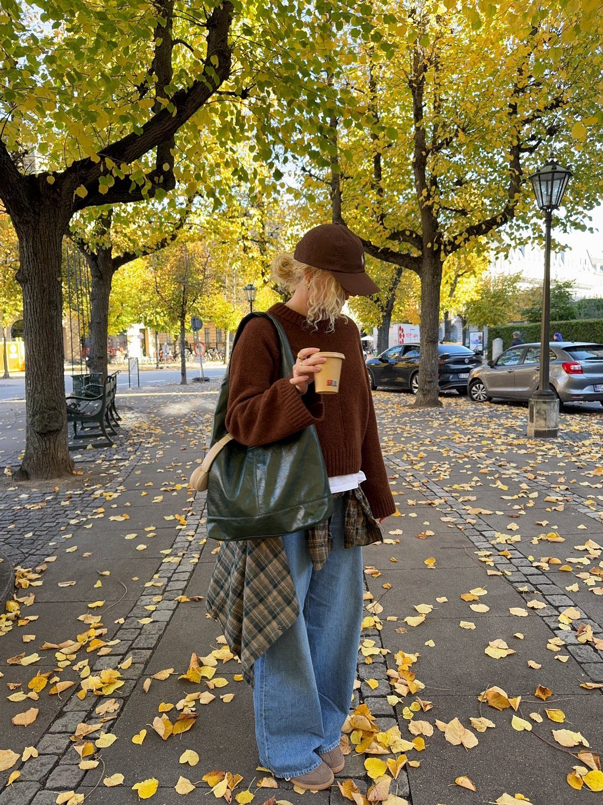 Woman in a brown sweater and wide-leg jeans walking through autumn leaves on a cobblestone path.