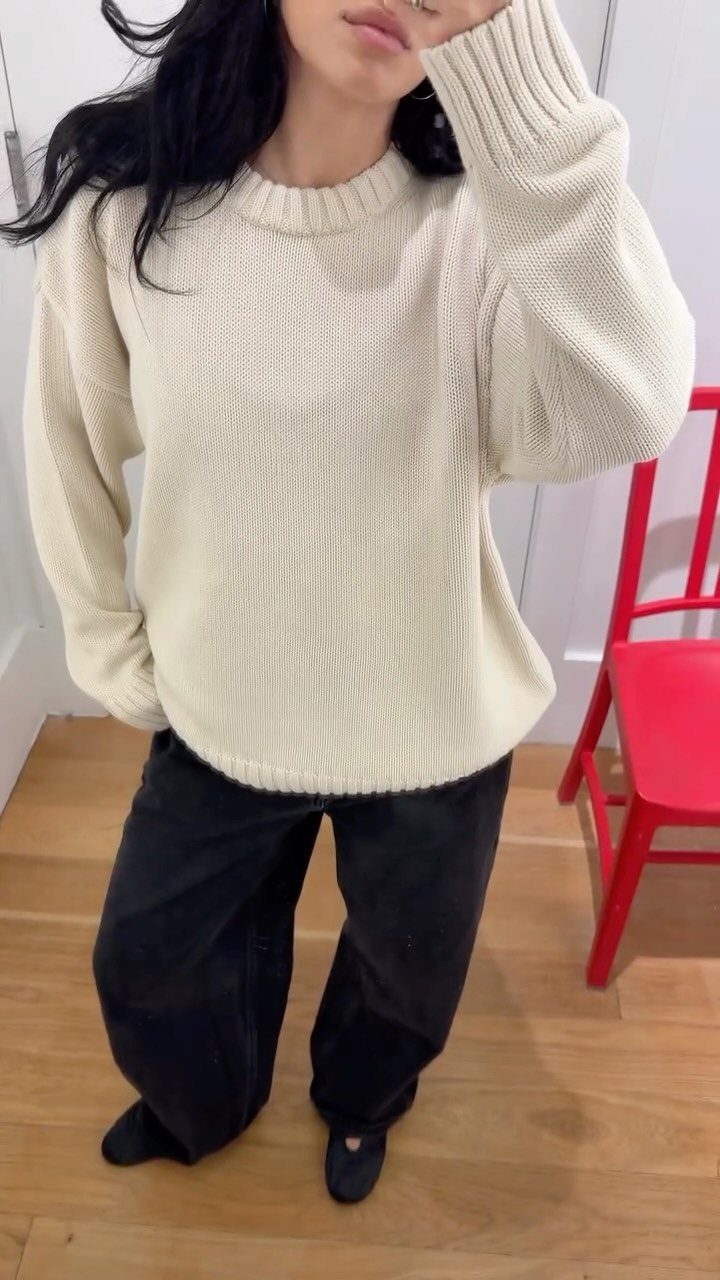Woman wearing a cream ribbed knit sweater and wide-leg black trousers standing indoors.