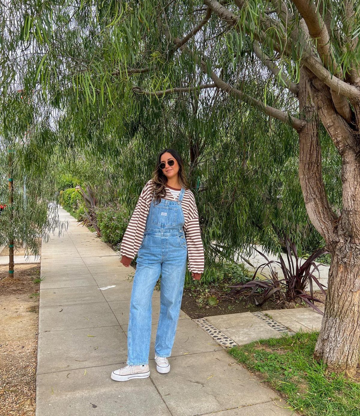 Woman in striped sweater and denim overalls standing on a sidewalk near trees.