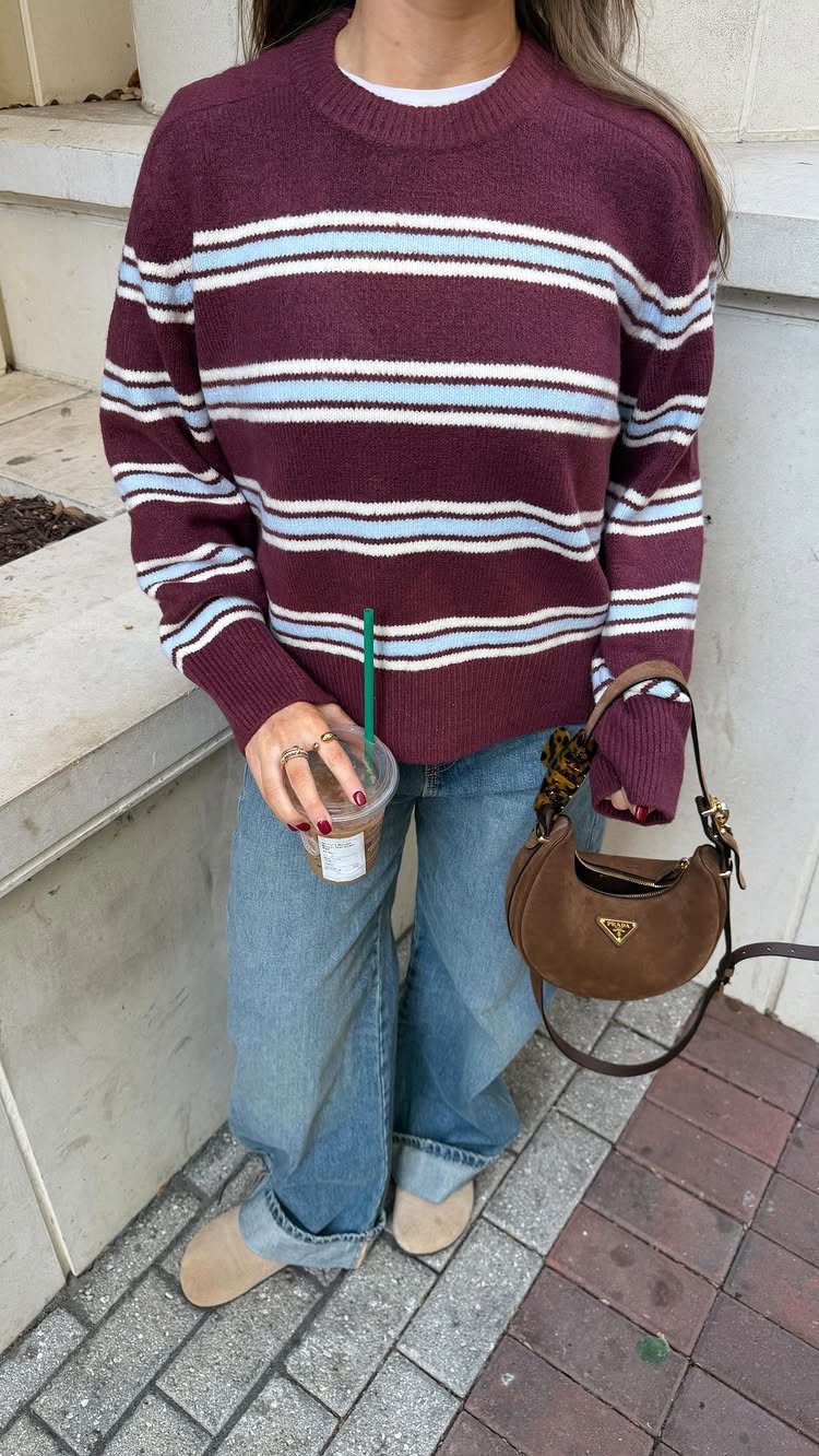A woman wearing a maroon striped sweater and light blue wide-leg jeans while holding a coffee.