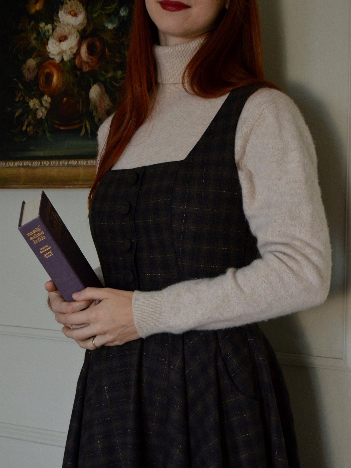 Person wearing a beige turtleneck under a dark grey plaid pinafore dress holding a book