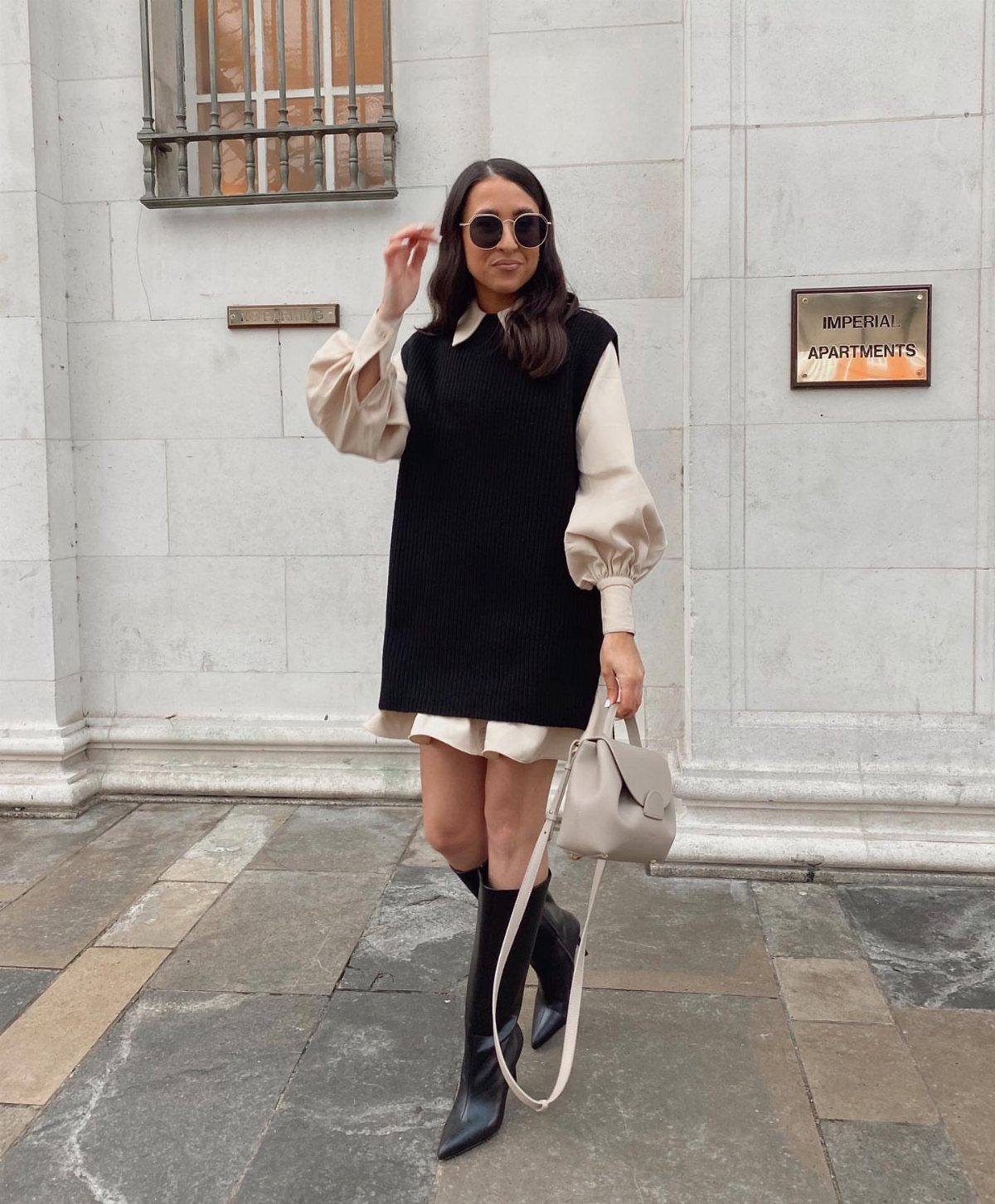 Woman wearing a black knit vest over a cream long-sleeved dress and black boots.