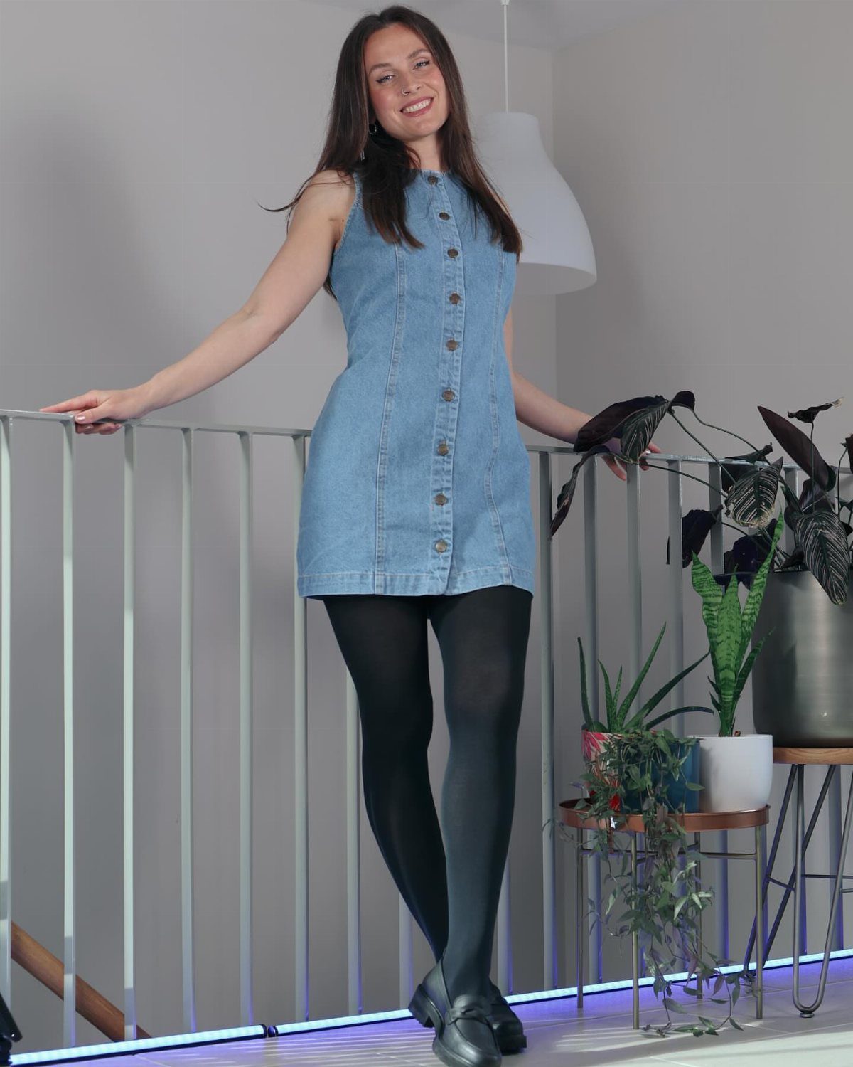 Woman wearing a sleeveless denim dress with black tights and black shoes indoors.