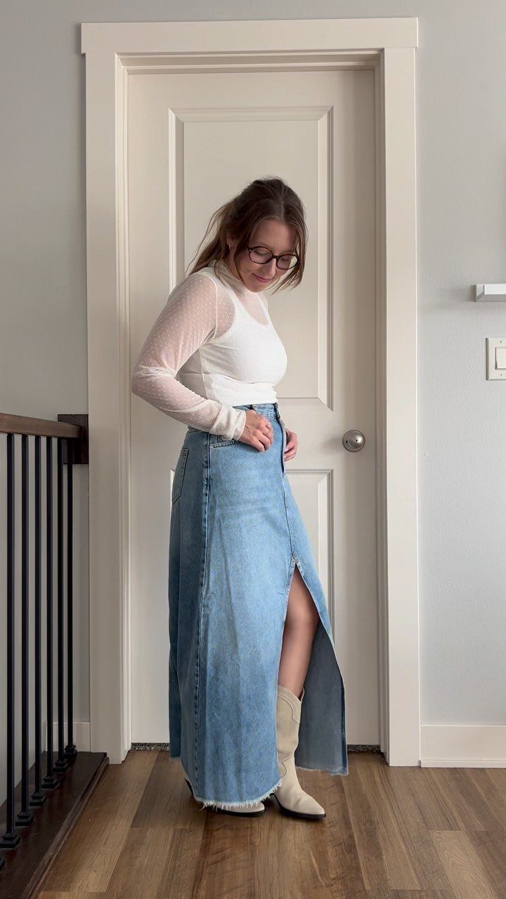 A woman posing in a white sheer top and a long denim maxi skirt with a slit.