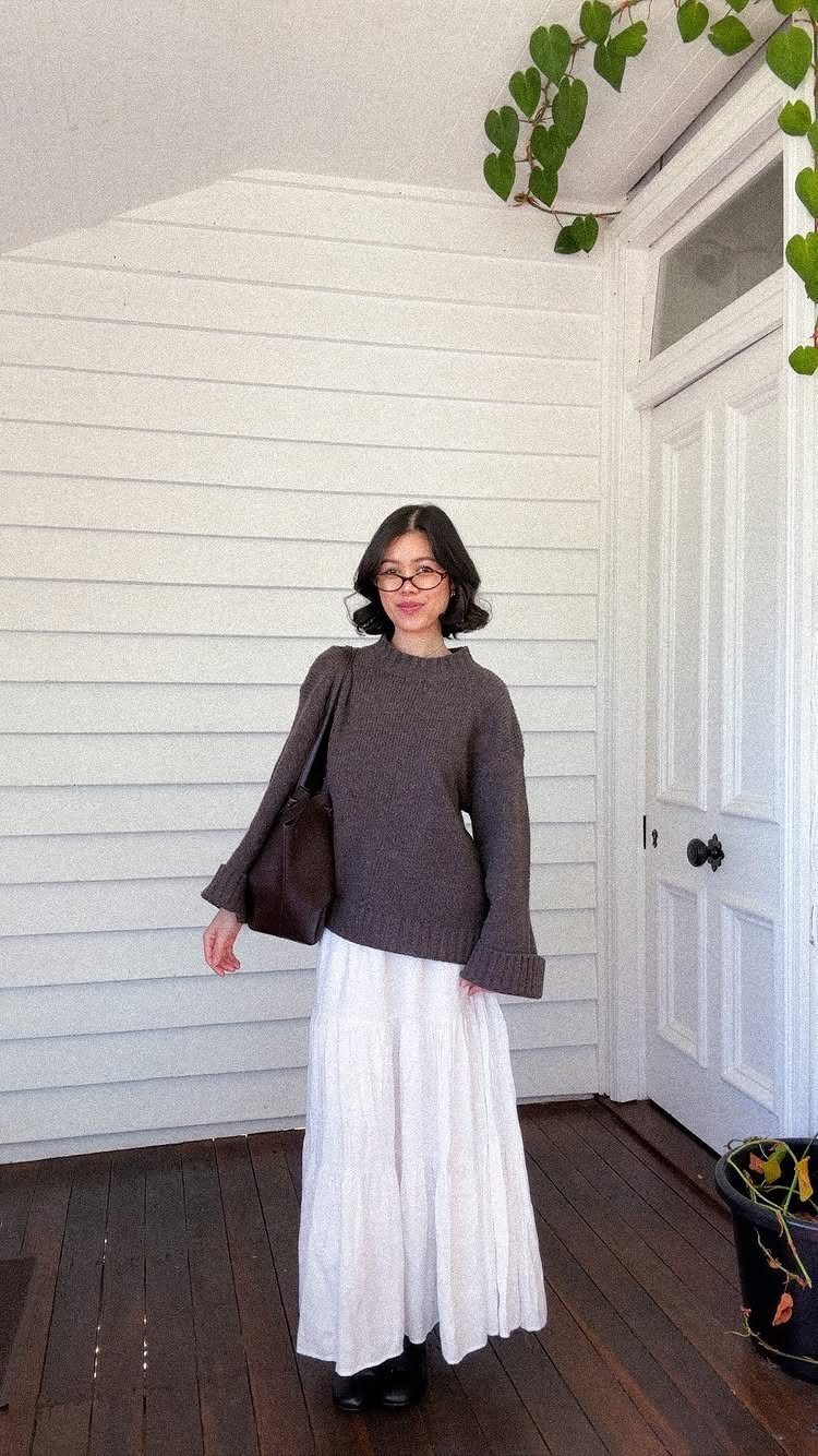 A woman wearing a brown sweater and a long white tiered skirt standing by a white door.