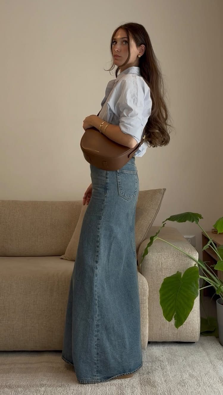 Woman wearing a light blue shirt and a long denim maxi skirt standing in a living room.