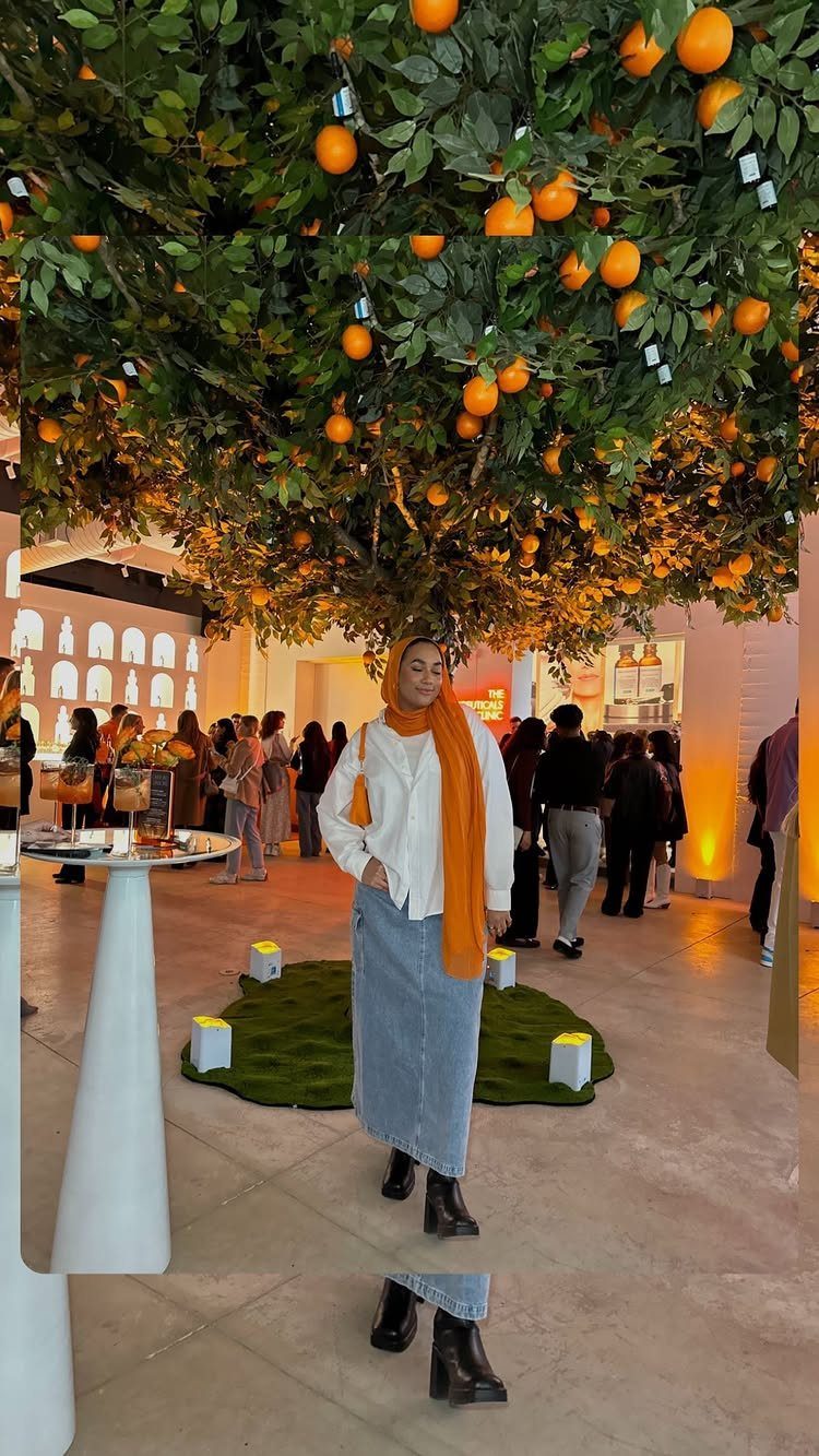 Woman wearing a white button-down shirt, long denim skirt, and orange scarf under orange trees.
