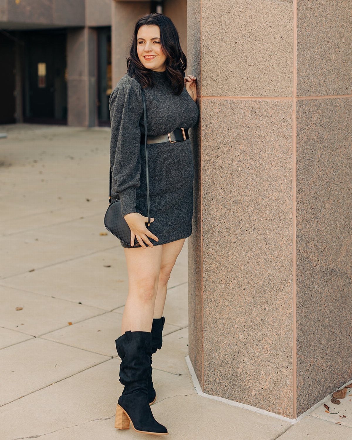 Woman in a grey sweater dress with a black belt and black boots leaning against a pillar.