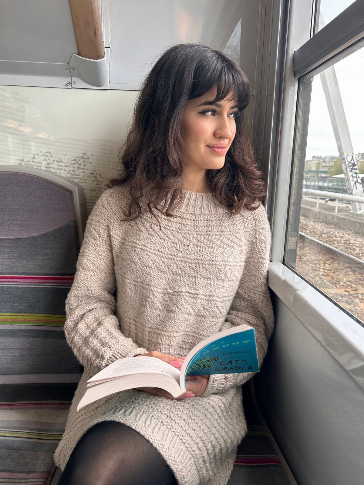 A woman in a beige knit sweater dress sitting on a train reading a book.