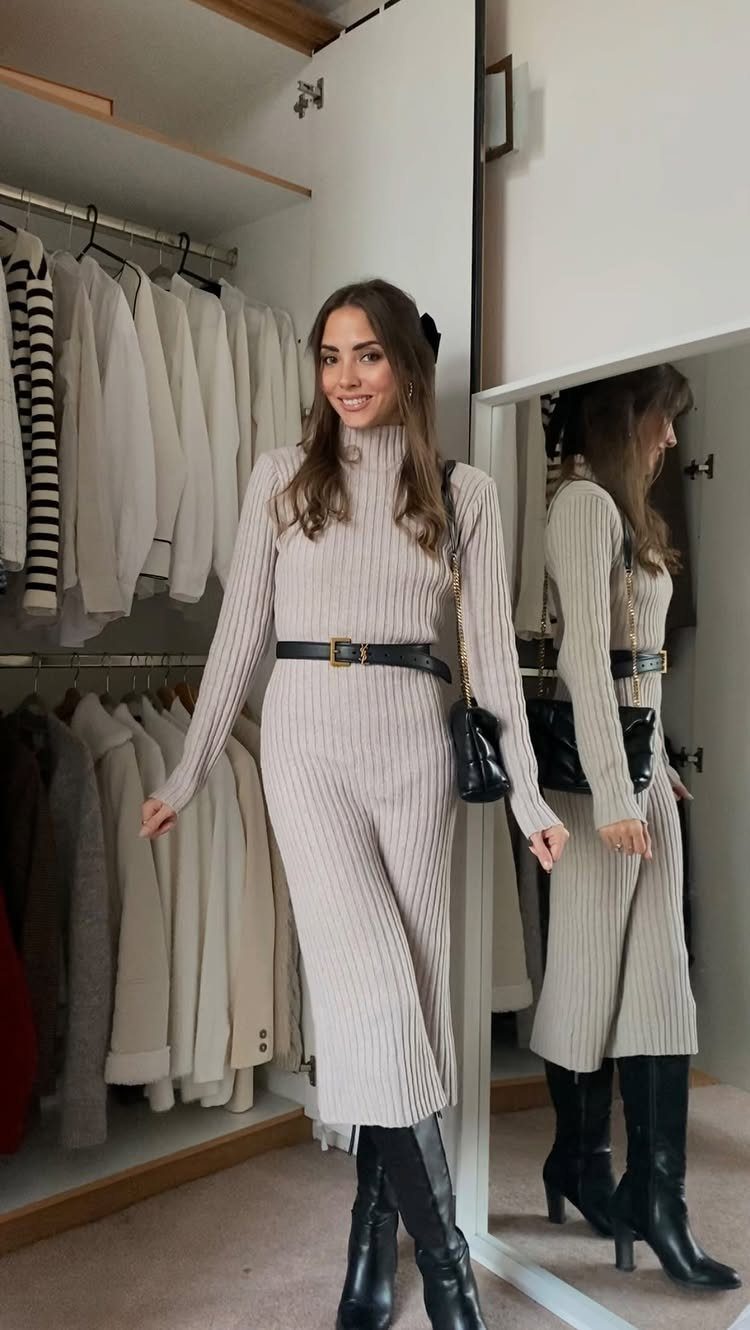 Woman in a beige ribbed sweater dress, black belt, and black knee-high boots posing in a closet.