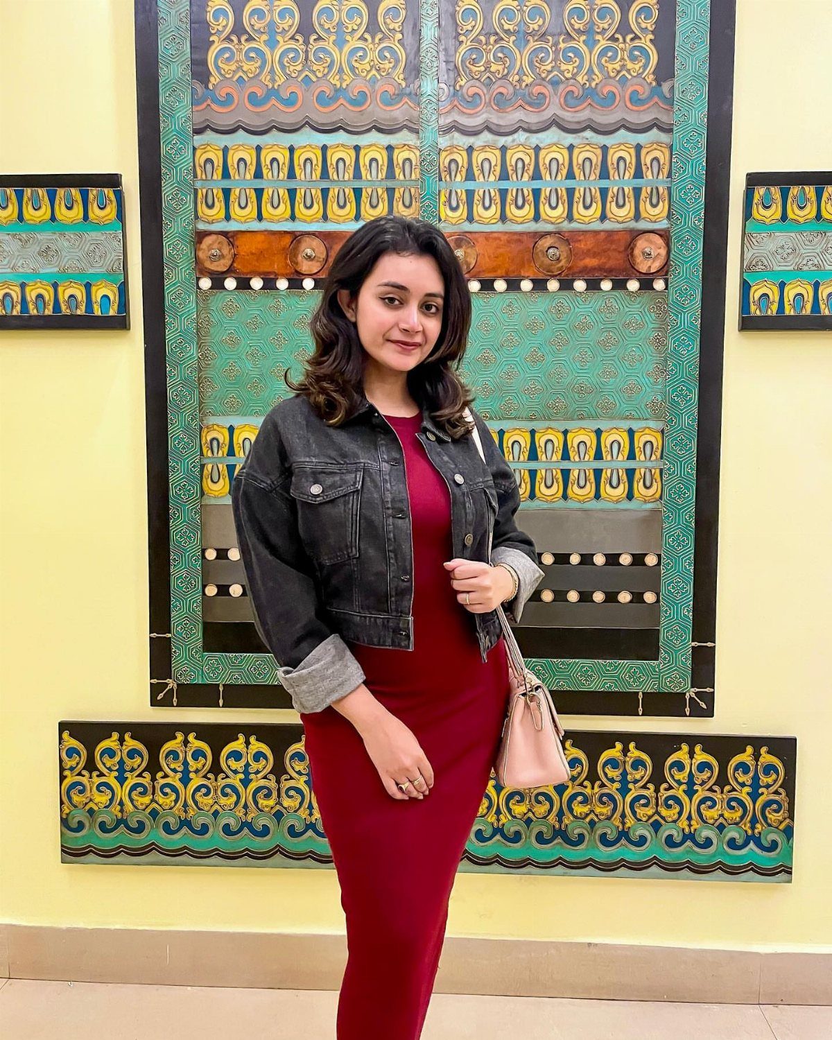 A woman in a burgundy sweater dress and black denim jacket posing against a patterned wall.