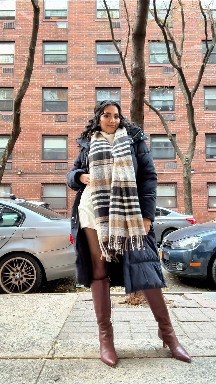 A woman wearing a cream sweater dress, dark coat, plaid scarf, and tall brown boots.