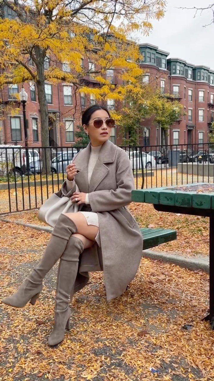 Woman in a cream sweater dress and grey over-the-knee boots sitting on a park bench.