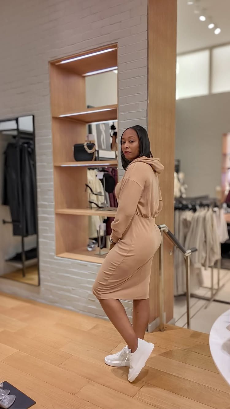 Woman in a tan hooded sweater dress and white sneakers posing in a boutique.
