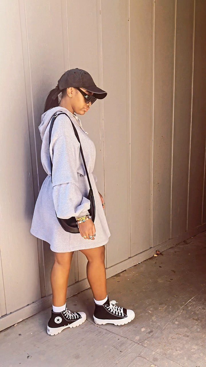 A woman in a grey hooded sweater dress, black cap, and sunglasses standing against a wall.